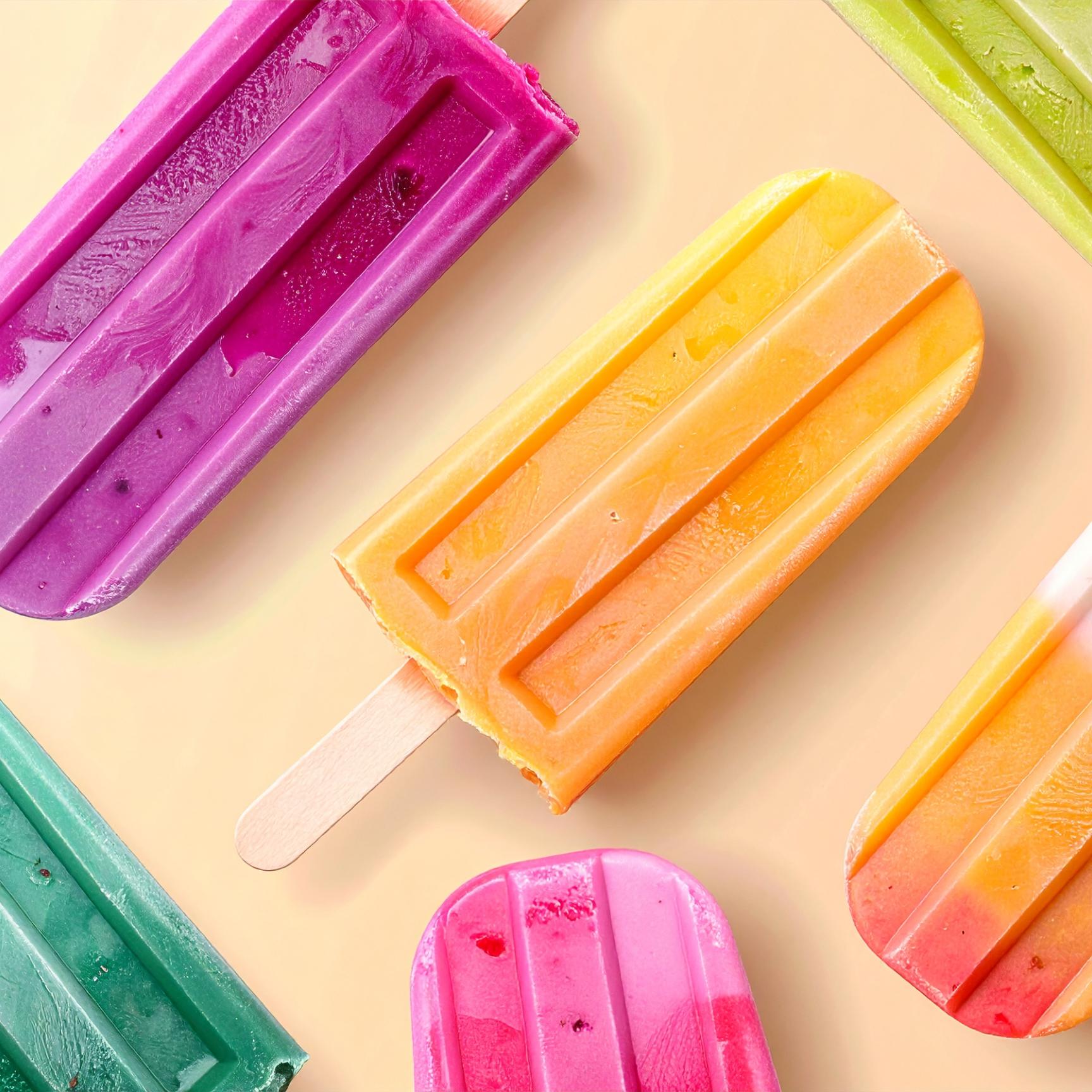 Sechs bunte Eis am Stiel liegen auf einer hellen Oberfläche, farbenfroh arrangiert in Lila, Orange, Grün, Pink und Rot – eines davon mit einem Biss. Die Szene wirkt sommerlich und einladend.
