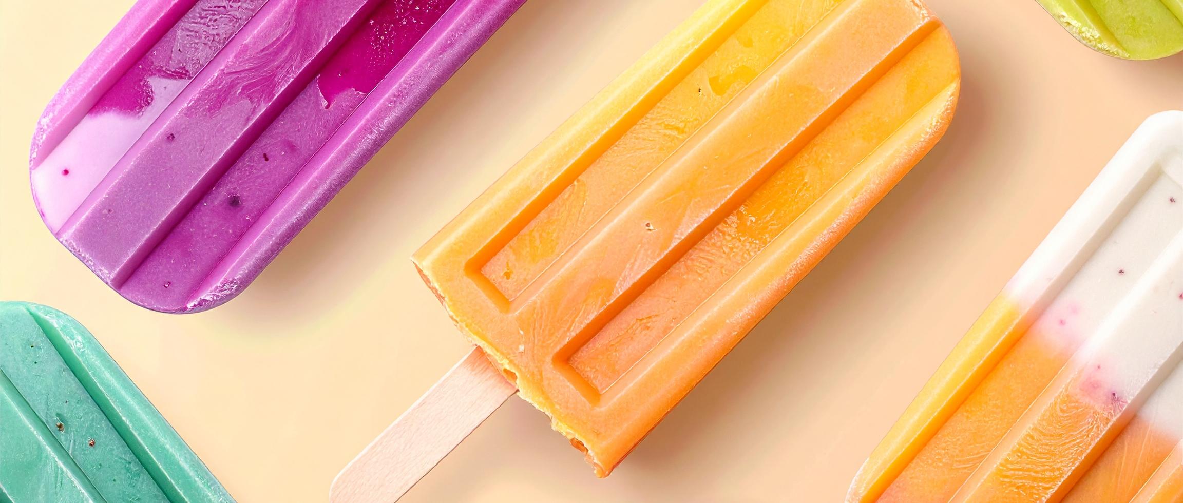 Sechs bunte Eis am Stiel liegen auf einer hellen Oberfläche, farbenfroh arrangiert in Lila, Orange, Grün, Pink und Rot – eines davon mit einem Biss. Die Szene wirkt sommerlich und einladend.