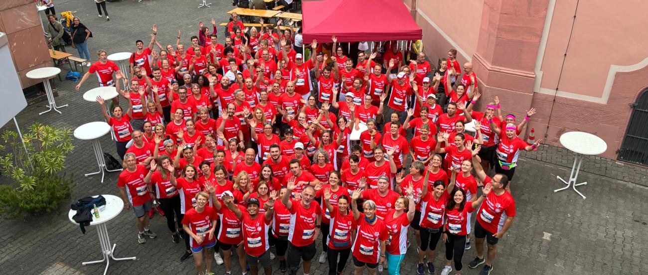 Eine große fröhliche Menschenmenge in roten T‑Shirts steht in einem Innenhof zusammen, winkt in die Kamera.