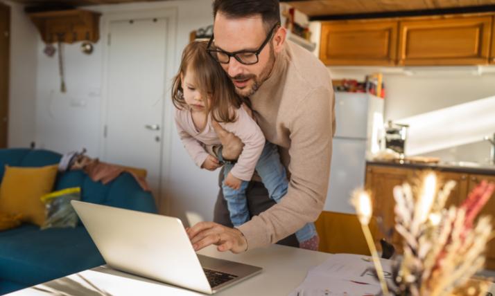 Vater mit Kind auf dem Arm arbeitet am Computer im Stehen