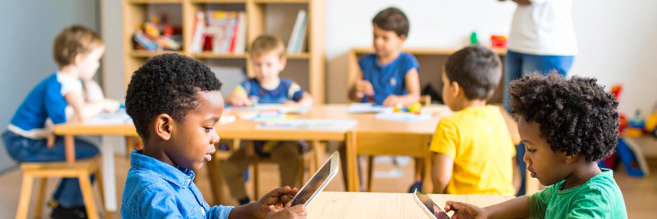 Mehrere Kinder sitzen in einem Kita-Raum an Tischen und nutzen Tablets. Im Hintergrund steht eine erwachsene Person, daneben ein Regal mit Büchern und Spielzeug.