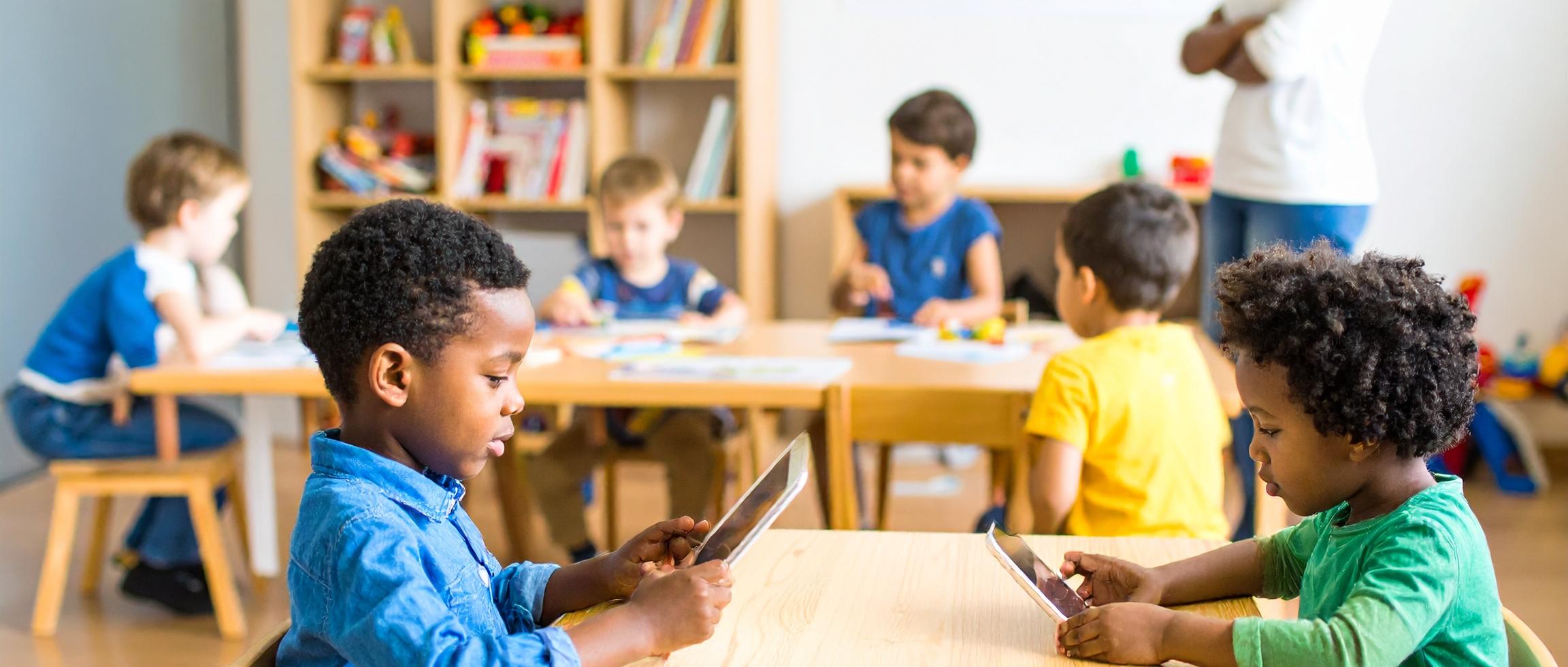 Mehrere Kinder sitzen in einem Kita-Raum an Tischen und nutzen Tablets. Im Hintergrund steht eine erwachsene Person, daneben ein Regal mit Büchern und Spielzeug.