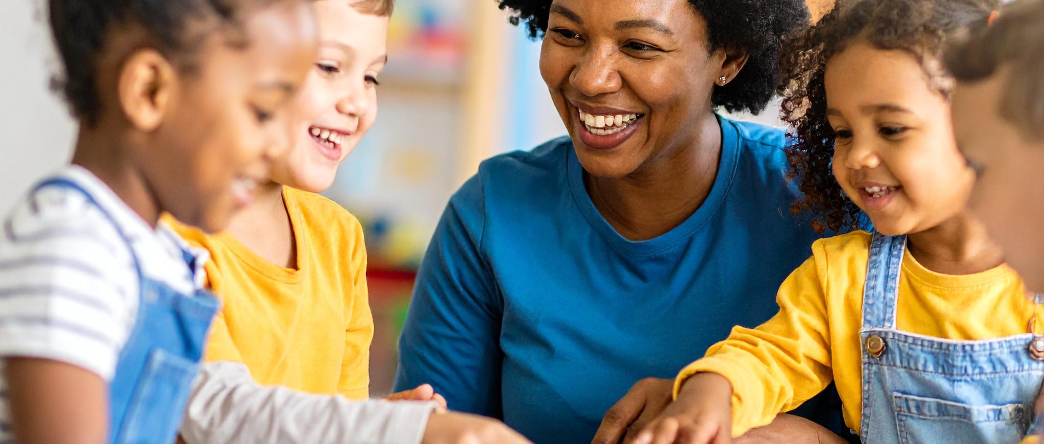 Eine Gruppe von Kindern spielt gemeinsam mit bunten Bauklötzen an einem Tisch, begleitet von einer erwachsenen Person in einem kindgerecht eingerichteten Raum. Die fröhliche Szene vermittelt einen Einblick in den pädagogischen Alltag einer Kindertagesstätte.