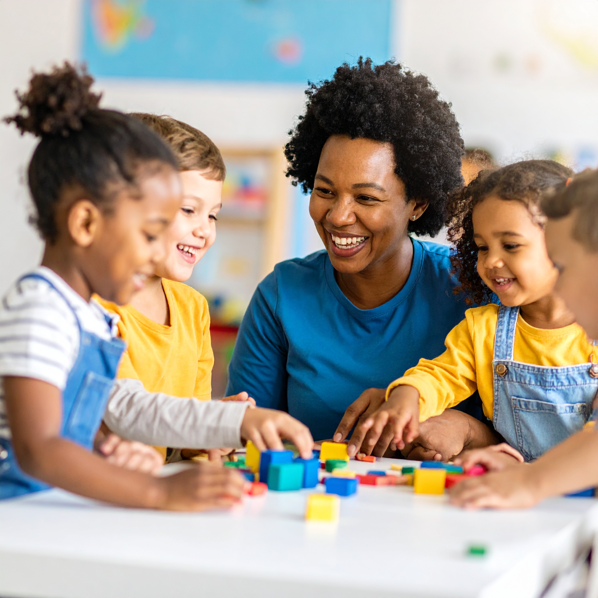 Eine Gruppe von Kindern spielt gemeinsam mit bunten Bauklötzen an einem Tisch, begleitet von einer erwachsenen Person in einem kindgerecht eingerichteten Raum. Die fröhliche Szene vermittelt einen Einblick in den pädagogischen Alltag einer Kindertagesstätte.