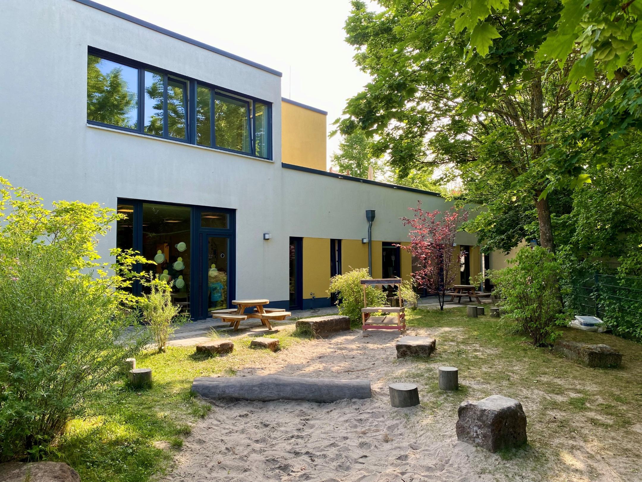 Gebäude von Außen, Sandkasten, Sitzbänke, Sonnenschein, begrüntes Außengelände
