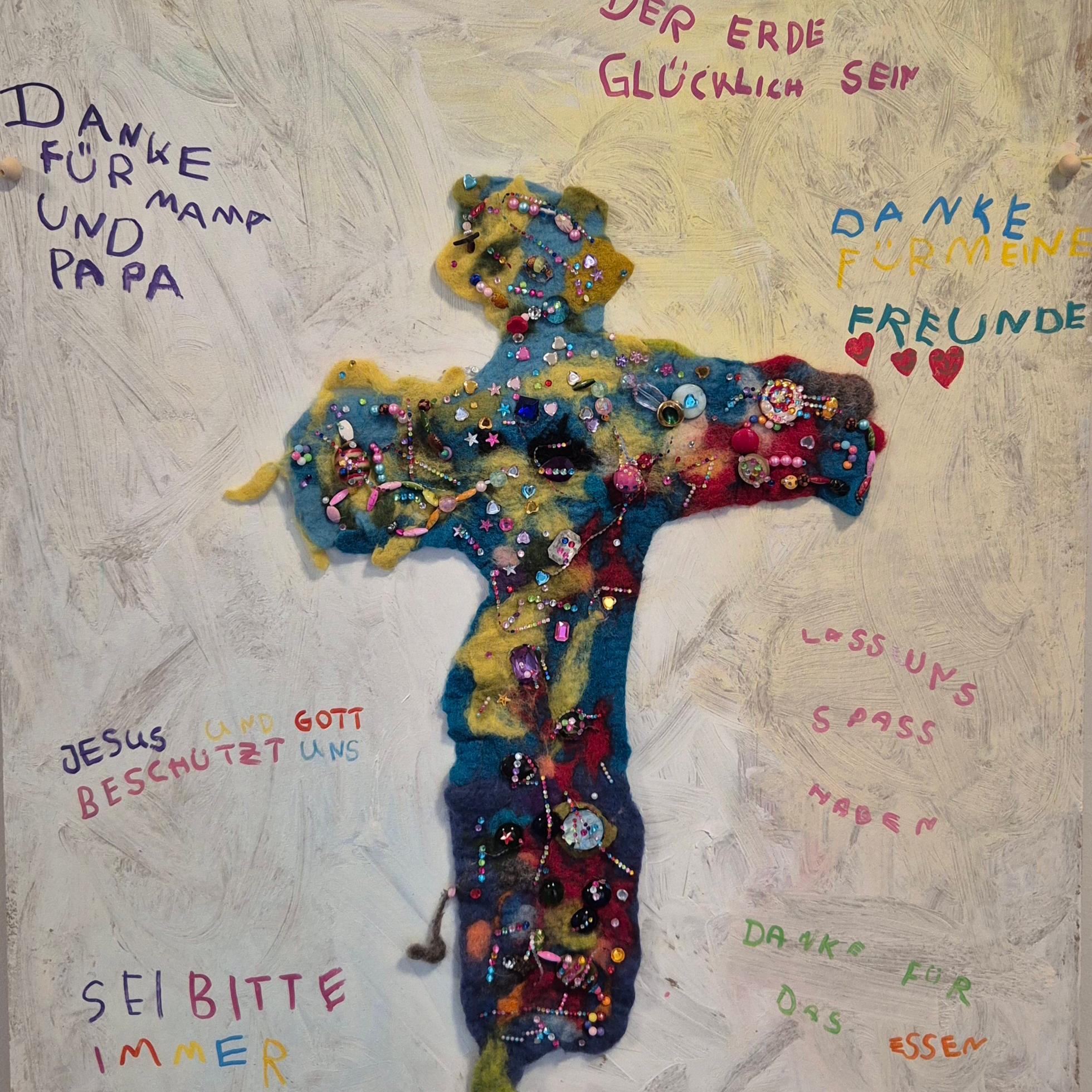 Ein gefilztes Jesuskreuz verziert mit bunten Perlen. Umrahmt mit kurzen Sätzen und Botschaften von Kindern